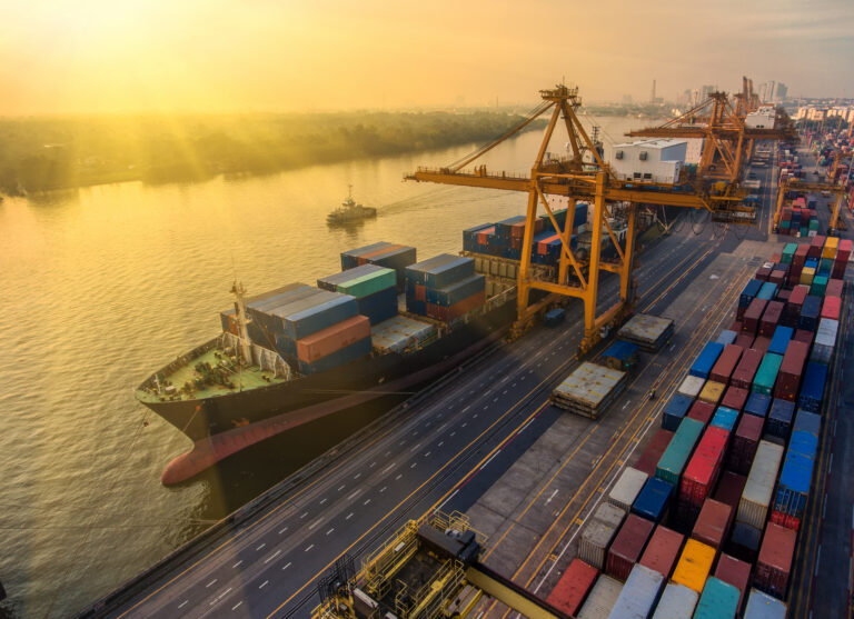 Container cargo ship being loaded at a commercial shipping port with cranes and stacked freight containers along the waterfront at sunrise.